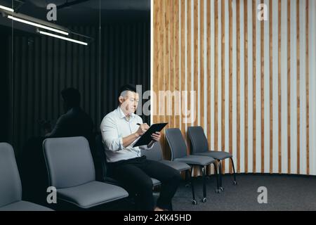 Ein junger gutaussehender Asiate sitzt ernsthaft in der Lobby des Bürozentrums, wartet auf ein Interview und hält einen Ordner mit Papieren und Dokumenten in seinen Händen. Er füllt gerade einen Fragebogen aus. Stockfoto