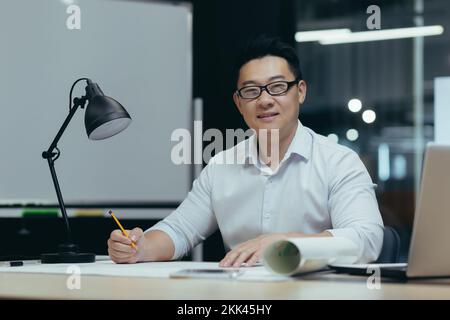 Porträt eines jungen, gutaussehenden Asiaten, Architekt, Designer, Freiberufler. Sie saßen im Büro am Tisch in einer Brille mit einem Laptop. Macht eine Zeichnung, macht einen Plan. Er sieht in die Kamera und lächelt. Stockfoto