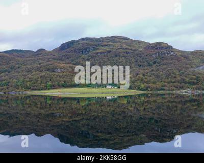 Haus eingebettet in Grasgebiet, umgeben von Hügeln, zerklüfteten Gebieten und Bäumen, perfekt reflektiert im Spiegelwasser von Loch Goil, Schottland, Großbritannien. Stockfoto