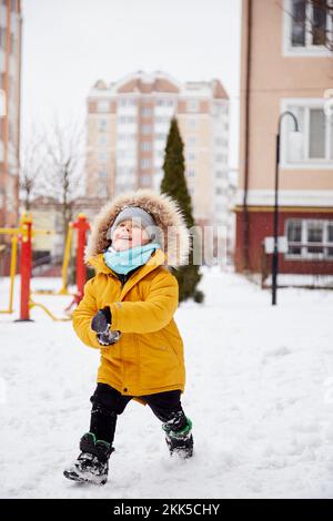 Glückliches Kind, das den Schnee im Hof der Stadt genießt. Winterspaß draußen. Junge in leuchtend orangefarbener Winterjacke. Stockfoto