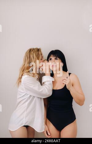 Eine aufgeregte Frau in einem schwarzen Badeanzug hört einer Freundin in einem Hemd zu, die ihr auf einem hellen Hintergrund ins Ohr flüstert Stockfoto