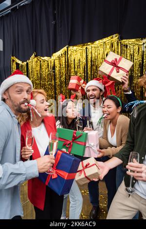 Fröhliche, gemischtrassige Geschäftsleute mit Partyhörnern, die im Büro Geschenke und Champagner halten Stockfoto