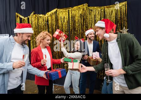 Lächelnde, multiethnische Geschäftsleute in weihnachtsmannhüten, die im Büro Champagner und Geschenke halten Stockfoto