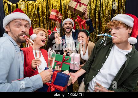 Lächelnde, multiethnische Geschäftsleute in weihnachtsmannhüten, die während der Party im Büro Champagner und Geschenke in der Nähe von Lametta hielten Stockfoto