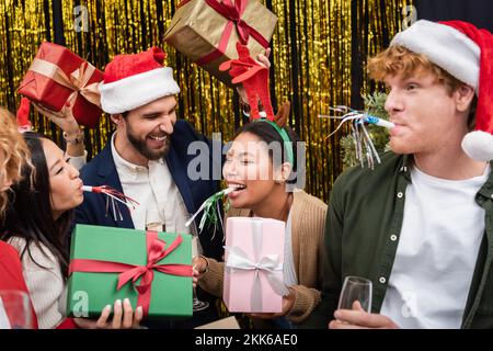 Multiethnische Geschäftsfrau im Weihnachtskopfband, die ein Geschenk hält und neben Kollegen im Büro ein Partyhorn bläst Stockfoto