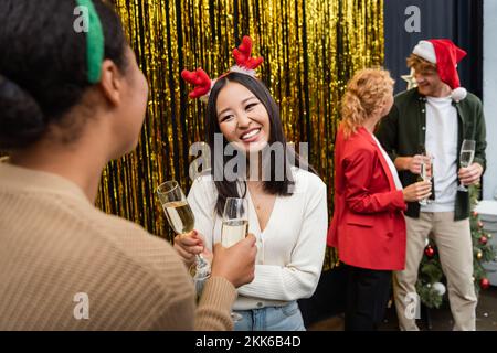 Lächelnde asiatische Geschäftsfrau, die Champagner hält und während der Weihnachtsfeier im Amt mit einem multiethnischen Kollegen spricht Stockfoto