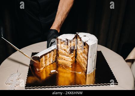 Fantastische Kuchen. Ein Koch mit schwarzen Handschuhen schneidet eine Schokoladenkuchen-Hochzeitstorte auf. Der Hochzeitskuchen ist innen köstlich auf schwarzem Hintergrund. Großer Kuchen in Weiß Stockfoto