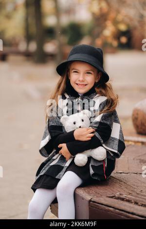 Ein kleines stylisches Mädchen in Herbstkleidung sitzt auf einer Bank in der Herbststadt Stockfoto