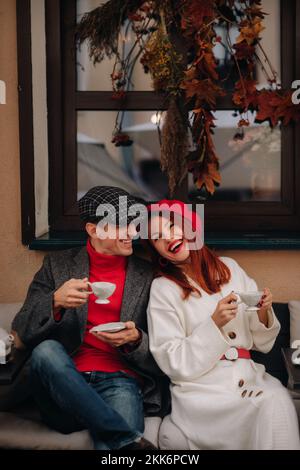 Ein glückliches, stilvolles Paar trinkt Kaffee und lächelt, während es in einem Café auf der Straße sitzt Stockfoto