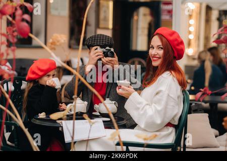 Eine stilvolle dreiköpfige Familie sitzt draußen an einem Tisch in einem Café und trinkt Kaffee. Papa fotografiert Mama in der Herbststadt Stockfoto