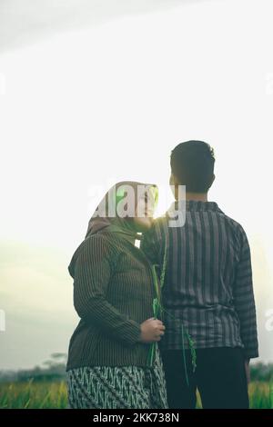 Ein paar Asiaten stehen Seite an Seite in traditioneller javanesischer Kleidung auf einem Reisfeld mit einem Hintergrund des Himmels. Fotoshooting vor der Hochzeit Stockfoto