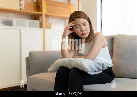 Eine junge asiatische Frau, die sich deprimiert und unglücklich fühlt und zu Hause auf einer Couch sitzt Stockfoto