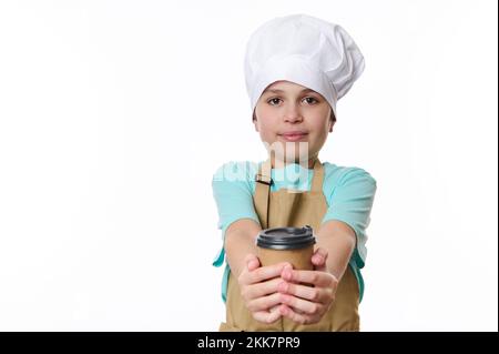 Ein multiethnischer, reizender Junge mit beigefarbener Schürze und weißer Kochmütze, der Kaffee zum Mitnehmen in einer Papiertasse anbietet Stockfoto