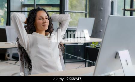 Entspannte, zufriedene junge Frau, die am Arbeitsplatz sitzt und ihre Arbeit genießt, glückliche, ruhige Geschäftsfrau, die sich in den Stuhl lehnt und die Hände hinter dem Kopf hält Stockfoto