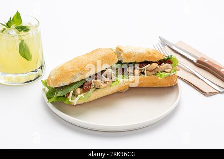 Ein halbgeschnittenes Sandwich mit Fleisch, Käse und Salat auf einer weißen Keramikplatte, mit Limonade in einem Glas und Besteck auf einer Serviette im Hintergrund Stockfoto