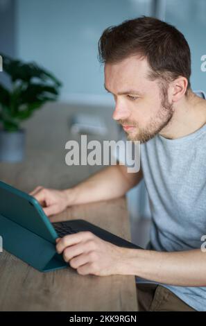 Fröhlicher Freiberufler mit Bart, der im Home Office sitzt und ein Tablet verwendet, um online zu arbeiten oder zu lernen. Nahaufnahme des Porträts eines jungen, gutaussehenden IT-Typen oder Ingenieurs in grauem Hemd. Hochwertiges vertikales Foto Stockfoto