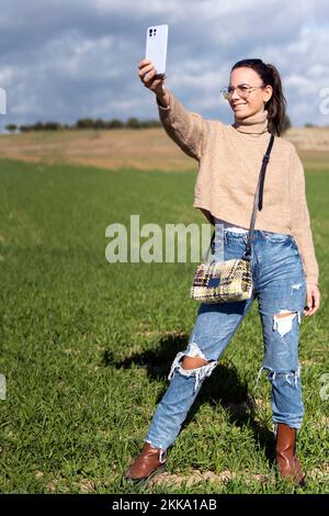 Fröhliche junge Frau in beigefarbenen Pullover-Jeans und Brille, die lächelt und Selfie auf dem Smartphone macht, während sie auf einer grünen Wiese steht Stockfoto