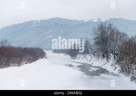 Moldawien, Rumänien, 2021-12-29. Gefrorener und schneebedeckter Fluss in Bucovina mit Nadelgebirgen im Hintergrund. Stockfoto