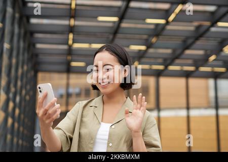 Glückliches asiatisches Mädchen winkt auf ihre Smartphone-Kamera, sagt Hallo zu einer Freundin im Video-Chat, ruft jemanden an, steht auf der Straße Stockfoto