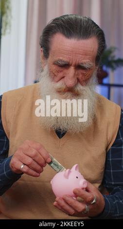 Der arme ältere Großvater hatte zu Hause nicht genug Geld. Er hatte eine Piggybank und eine Dollar-Banknote. Finanzkrise. Bankrott. Armut und Elend. Alter Mann auf der Couch. Vertikaler Schuss Stockfoto