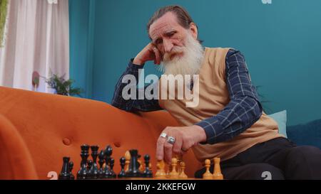 Fokussierter Senior Großvater Mann Schachmann sitzt zu Hause Raum Couch lernen spielen Schachbrett Spiel allein, pädagogische Freizeit häusliche Tätigkeit, strategische Fähigkeiten. Älterer Rentner Stockfoto