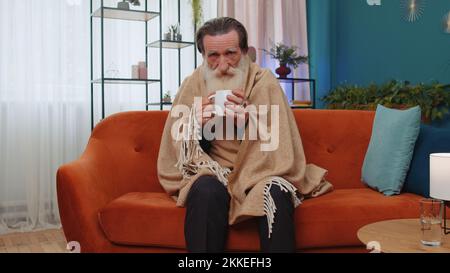 Kranker älterer Großvater, eingewickelt in karierte Form, zitterte vor Kälte auf dem Sofa und trank heißen Tee in einer unbeheizten Wohnung ohne Heizung aufgrund von Schulden. Ungesunde ältere Opa Gefühl Unbehagen versuchen, sich aufzuwärmen Stockfoto