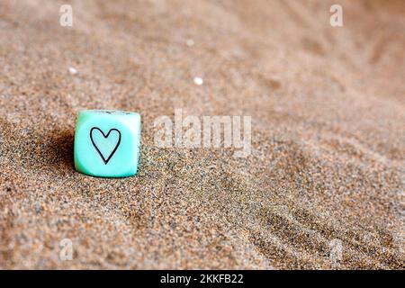 hellblauer Würfel mit Herz- und Wettersymbolen an den Seiten. Lustige Spielwürfel auf Sand. Valentinstag-Konzept. Liebessymbol. Nahaufnahme mit Kopie Stockfoto