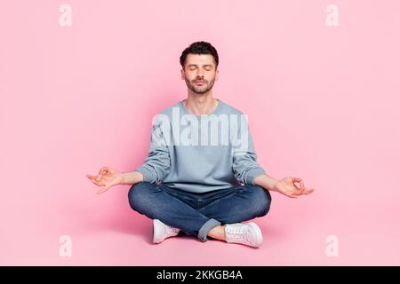 Ganzkörperfoto eines netten jungen Mannes, der auf dem Boden sitzt, einen entspannenden Arbeiter meditiert, der trendiges blaues Kleidungsstück gekleidet hat, isoliert auf pinkfarbenem Hintergrund Stockfoto