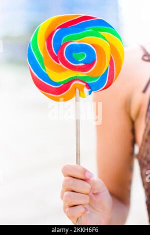Eine lebendige und farbenfrohe große Candy-Lollipop durch die Hand einer Frau vor dem Strand Hintergrund als Bild der Kindheit statt Stockfoto