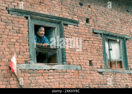 Ein altes Weibchen, das aus einem offenen Fenster eines alten Backsteinhauses schaut Stockfoto