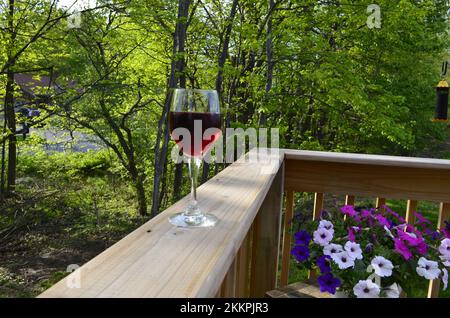 Ein volles Glas fruchtigen Rotwein auf einem hölzernen Geländer mit Blumen und Bäumen an einem wunderschönen Sommertag. Stockfoto