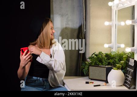 Frau im Spiegel der Garderobe kommuniziert per Videoanruf, hört Podcast, macht Selfie. Ein hübsches Mädchen trägt einen schwarzen Hut und lässige Kleidung hält die rote Mafia Stockfoto