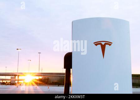 Ein rotes Tesla Logo auf der Vorderseite eines Tesla Supercharger ist zu sehen, wenn der Sonnenaufgang am Morgen im Hintergrund scheint. Stockfoto