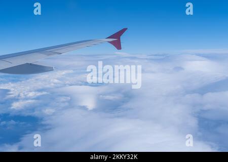 Der Blick vom Fenster des Flugzeugs auf den Flügel des Flugzeugs vor dem Hintergrund von flauschigen lockigen Wolken und blauem Himmel. Das Konzept des Reisens. Stockfoto