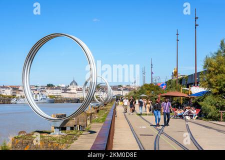 Die Menschen schlendern an einem sonnigen Tag auf der Insel Nantes, Frankreich, entlang des „Hangar à Bananes“-Gebäudes, gesäumt von „Les Anneaux“ von Buren und Straßencafés Stockfoto