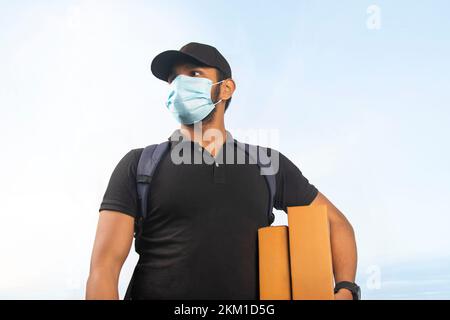 Portrait des jungen Liefermanns mit Gesichtsmaske und Pappkarton im Freien Stockfoto