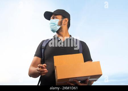 Portrait des jungen Liefermanns mit Gesichtsmaske und Pappkarton im Freien Stockfoto