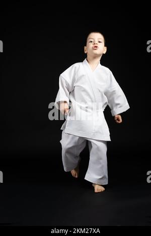 Der kleine starke Junge im Kimono geht voran, übt Karate, schwarzer Hintergrund Stockfoto