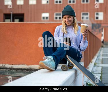 Nachhaltigkeit Lebensweise, junge Frau trinkt aus einer recycelbaren Tasse Kaffee, Skaterin hört Musik mit Smartphone, Longboard-Öko Stockfoto