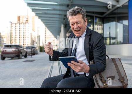 Geschäftsmann feiert Erfolg sieht auf Tablet-PC nach Erfolg aus, Senior Entrepreneur drückt positive Emotionen aus, sitzt draußen mit einem Wolkenkratzer im Büro Stockfoto