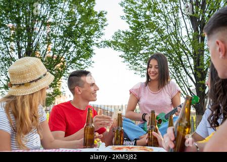 Leute, die Spaß an einem Holztisch mit Pizza zum Mitnehmen und Flaschen Bier haben, junge Paare, die singen und Musik spielen, um sich sorgenfrei zu fühlen Stockfoto