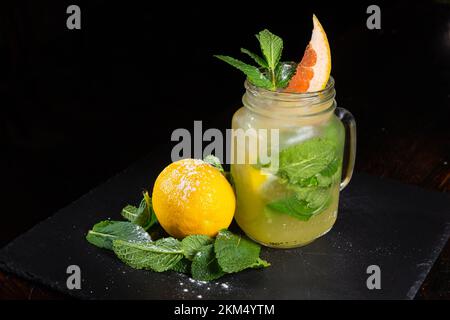 Mason-Glas mit leckerer kalter Limonade dekoriert mit Grapefruit, Zitrone und Minze auf schwarzem Hintergrund. Sommerentgiftungsgetränk mit frischem Obst im Glasbehälter. Stockfoto