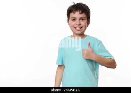 Ein glücklicher, weißer Junge zeigt einen Daumen nach oben und schaut in die Kamera, isoliert auf weißem Hintergrund. Kostenlose Werbefläche Stockfoto