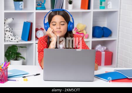 Müde und gelangweilte Teenager. Eine junge Studentin, die am Tisch sitzt und beim Lernen Kopfhörer und Laptop benutzt. Fernunterricht Stockfoto