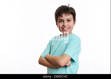 Ein reizender vorpubertär Junge, der vor der Kamera lächelt, mit einem schönen, zahnigen Lächeln, mit gekreuzten Armen auf weißem Hintergrund posiert Stockfoto