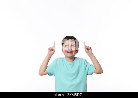 Porträt eines netten jungen Kindes in einem blauen lässigen T-Shirt, der mit dem Finger nach oben auf den Textbereich zeigt, auf weißem Hintergrund Stockfoto