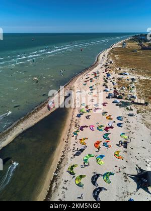 Blick von oben auf helle farbenfrohe Drachen, die am windigen Tag am Kitesurfplatz am Strand geparkt sind. Viele Fallschirme zum Kiteboarden liegen am Ufer Stockfoto