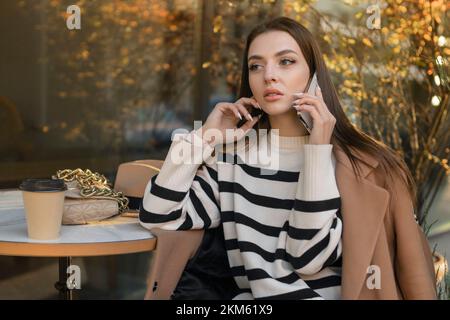 Eine brünette, wunderschöne, stilvolle, weiße Frau in lässiger Kleidung auf einem Spaziergang entlang der Straße einer europäischen Stadt sitzt in einem Café und spricht am Telefon Stockfoto