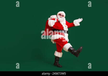 Ganzkörper-Profil Seitenfoto von santa claus mit groß Bauchbart tragen Weihnachtsgeschenk Geschenk Sack gehen tragen Kopfbedeckung Kappe Sonnenglas isoliert über hell Stockfoto