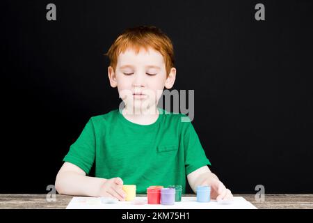 Porträt eines kleinen rothaarigen Jungen zeichnet mit Farben auf Papier, der Junge zeichnet mit seinen Händen und deshalb sind seine Hände gemalt i Stockfoto